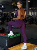 Picture of a model posing at the gym wearing our V back leggings in purple passion color along with the matching bra 