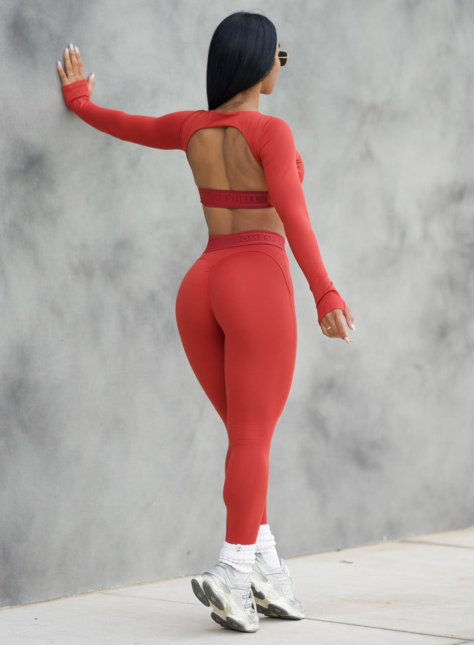 Model in outdoors using the wall as support with one hand and showing her back side profile wearing the Bombshell Active Leggings in Red ochre color