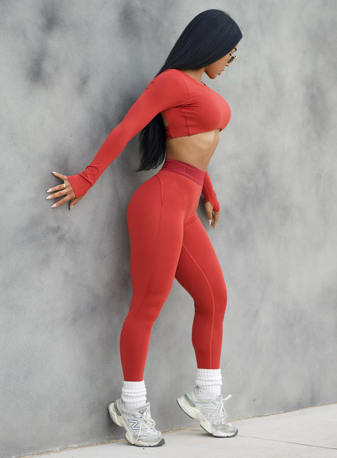 Model in outdoors leaning on a wall showing her right side profile wearing the Bombshell Active Leggings in Red ochre color