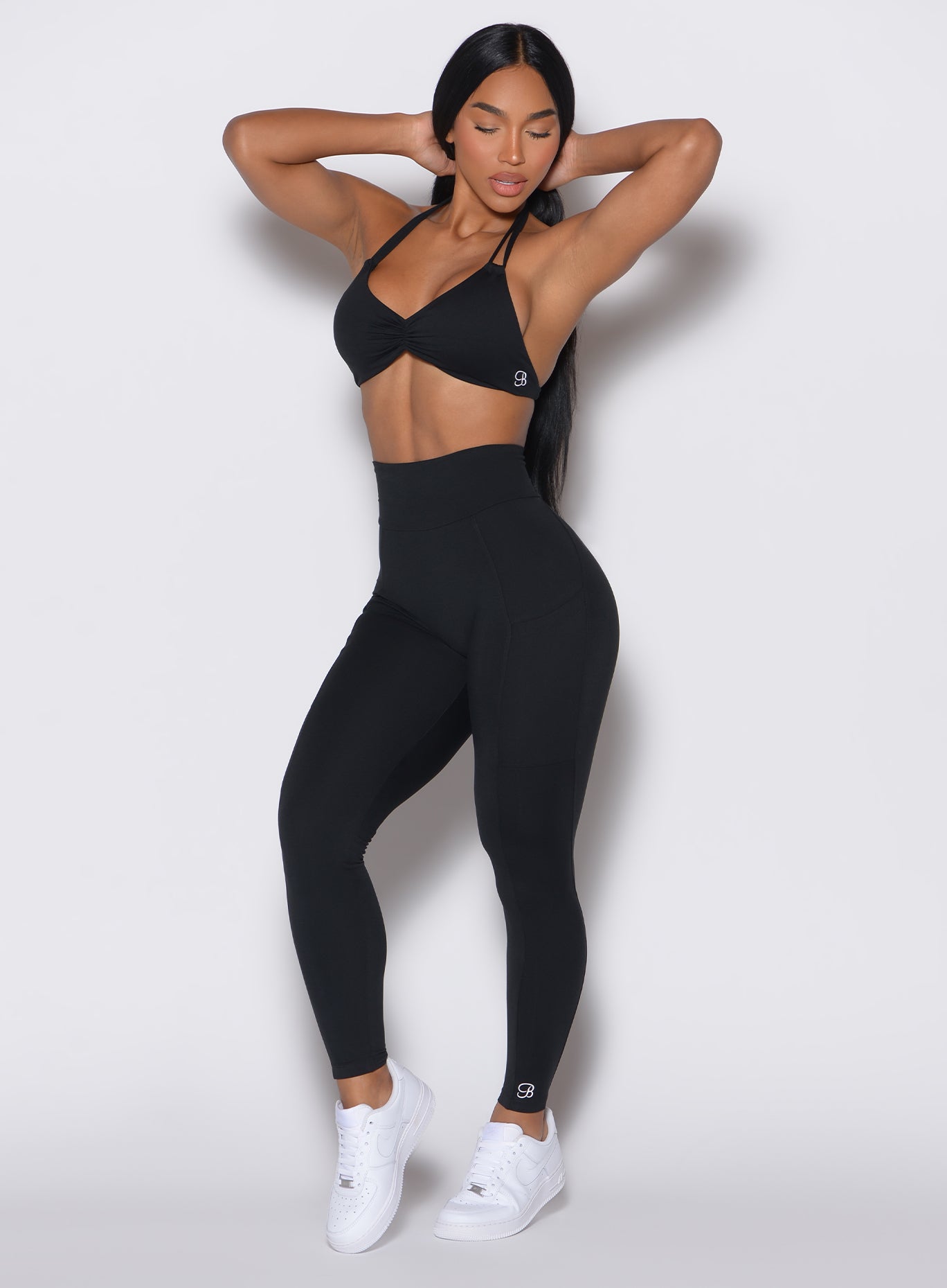 A front profile view captures a model elegantly posing with her hands behind her head, adorned in sleek black Curves leggings paired with a matching sports bra.