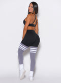 Back right side profile view of a model wearing the Perform Thigh Highs in Black and Smoke color with three white stripes on the thighs and a black sports bra