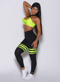 Left side profile view of a model angled to her left wearing our scrunch thigh high in black with neon yellow stripes on both thighs and a matching sports bra