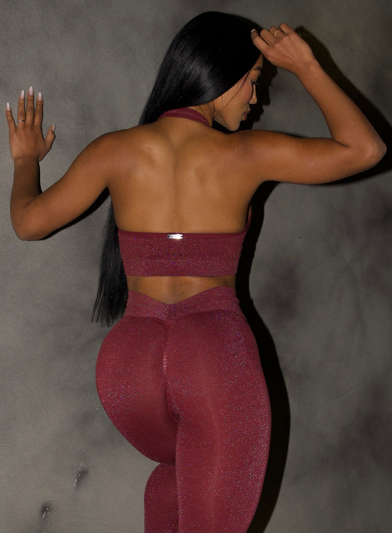 back profile view of a model with one hand on a wall wearing the Shimmer Backless Bra in Ruby star color