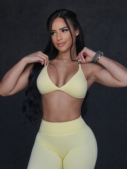 A woman with long, dark hair poses against a dark background wearing a pastel yellow athletic set. The set features a triangle bralette-style sports top and matching high-waisted leggings.