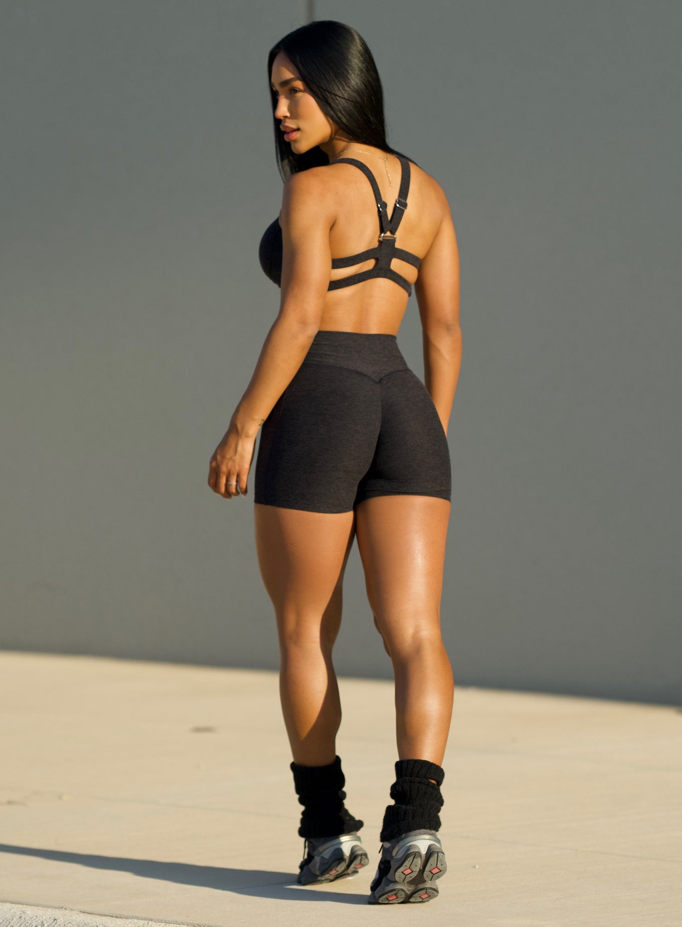 back profile of a model in outdoor sunshine wearing the Tiny Waist Shorts in Onyx color and a matching sports bra
