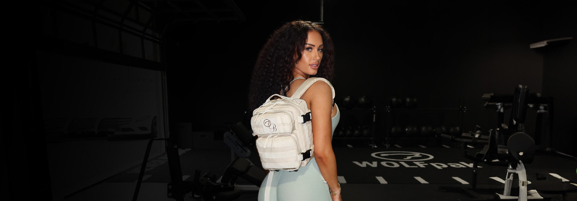 back view of model in gym wearing light blue bodysuit and cream small backpack