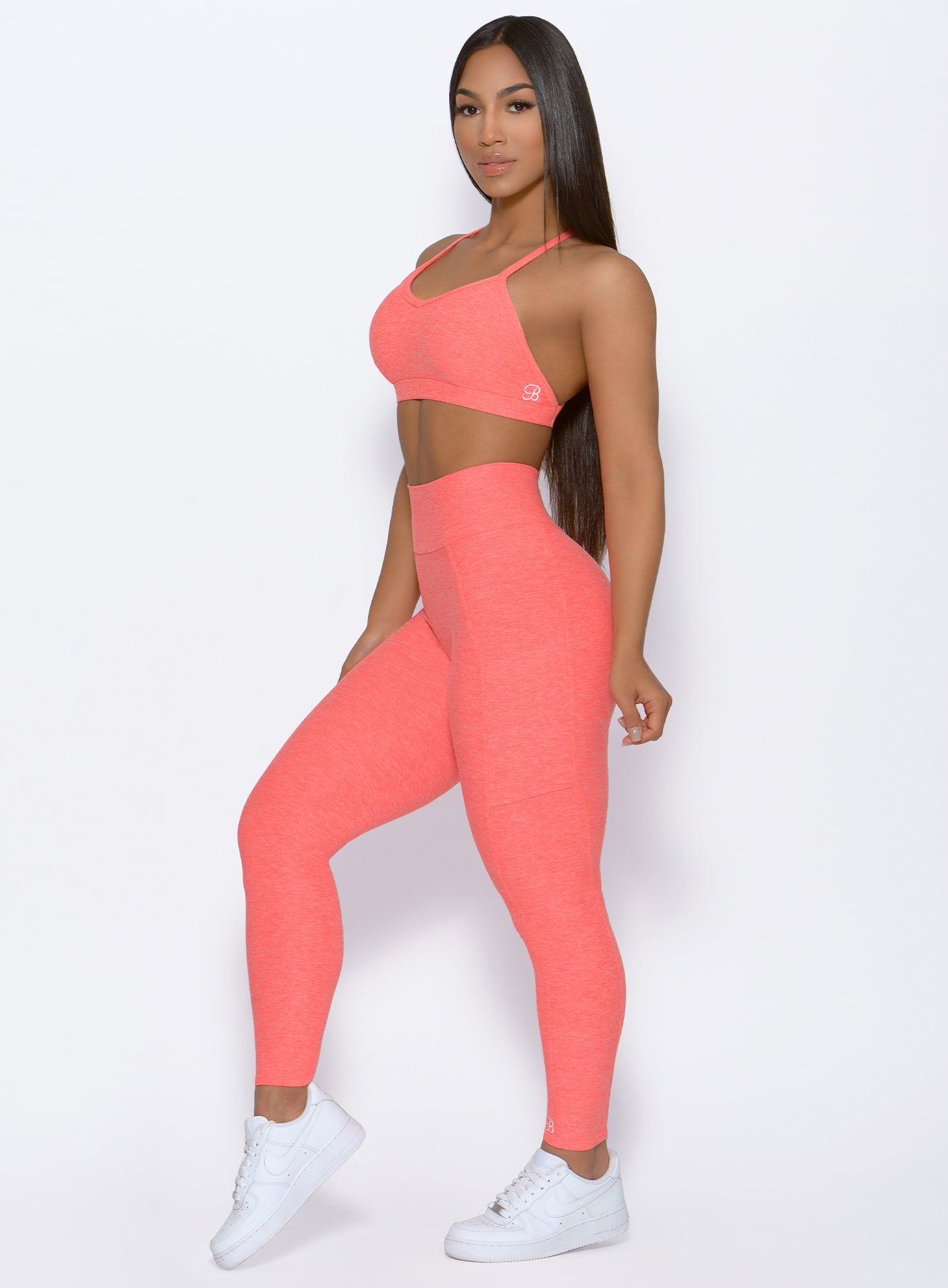 left side profile view of a model wearing our Curve leggings and the Braid Back Sports Bra in Coral color